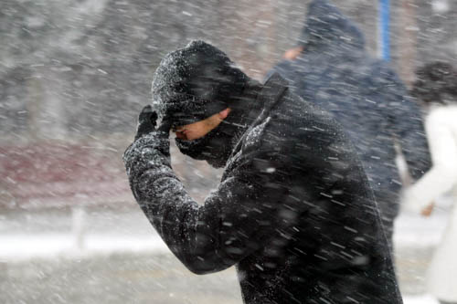 A man walks through a wild snowstorm holding a scarf to his face.